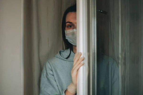 Kobieta w masce ochronnej i rękawiczkach wyglądająca przez okno domu z powodu epidemii wirusa korony covid-19. Koncepcja kwarantanny domowej i samoizolacji. — Zdjęcie stockowe