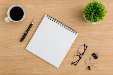 Üst Manzara Ahşap ofis masası. Ağaç demliği, defter, kahve fincanı, gözlük, kalem, ataç ofis malzemelerinin olduğu düz bir çalışma alanı.