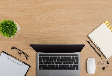 Üst Manzara Ahşap ofis masası. Düz çalışma alanı, gözlüklü, ağaç kabı, dizüstü bilgisayar, fare bilgisayarı, ataç, not panosu, defter, kalem, kalem ofis malzemeleri ahşap arka planda.