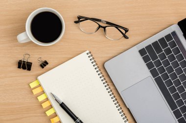 Üst Manzara Ahşap ofis masası. Kahve fincanı, dizüstü bilgisayar, defter, kalem, ataç ofis malzemeleriyle ahşap masa arkasında düz bir çalışma alanı.