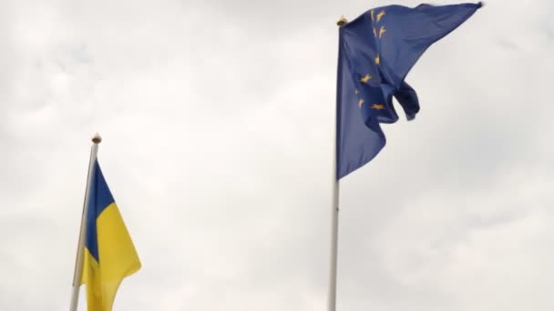 Fahne der Ukraine und Fahne der Europäischen Union flattern vor wolkenverhangenem Himmel.