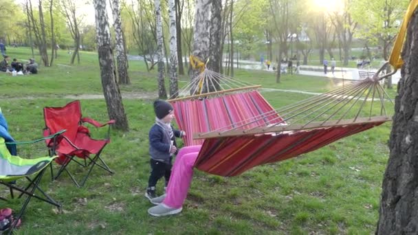 子供は 日没時に都市公園における色のハンモックで休みます 少年と少女が公園でハンモックで休んでいます — ストック動画