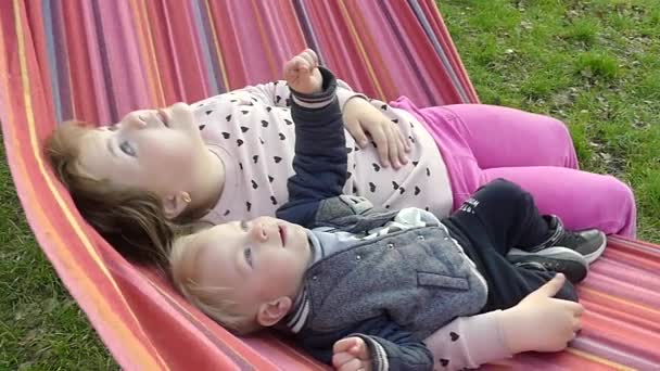 子供は 日没時に都市公園における色のハンモックで休みます 少年と少女が公園でハンモックで休んでいます — ストック動画