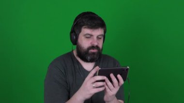 Bearded man in headphones communicates on Skype on a green background. A bearded man in headphones is talking via video link on a green chroma key. A man is laughing at a smartphone.