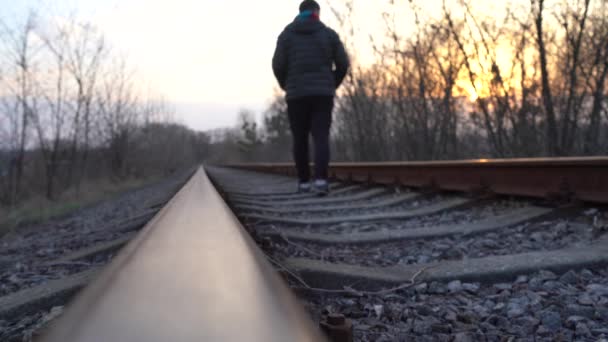 男が夕日に対して鉄道に沿って行きます 難民がレールを歩いている 電車の中で危険な散歩 — ストック動画