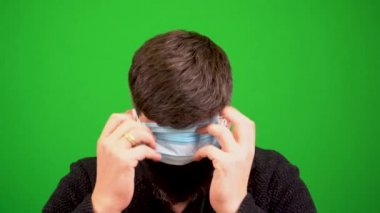 Sakallı bir adam koronavirüs salgını sırasında birçok tıbbi maske takar. Birçok tıbbi maske sakallı bir adama takılır. Bir adamın üzerinde bir kaç tane sargı bezi var. Yeşil arka planda bir yüzün yakın plan görüntüsü.