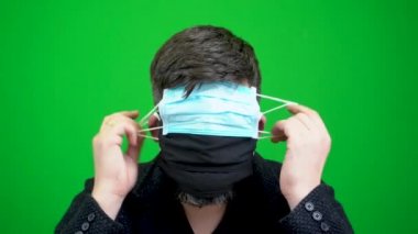 Sakallı bir adam koronavirüs salgını sırasında birçok tıbbi maske takar. Birçok tıbbi maske sakallı bir adama takılır. Bir adamın üzerinde bir kaç tane sargı bezi var. Yeşil arka planda bir yüzün yakın plan görüntüsü.