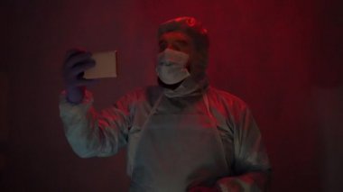 Laboratuvarda anksiyete sırasında koruyucu giysi giymiş sakallı bir doktor. Coronavirus salgını sırasında laboratuvarda bir kaza olmuş. Sirenin ışığı mavi-kırmızıdır. Odadaki kötü ışığın etkisi..