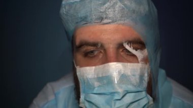 Yorgun sakallı bir doktor sisli bir maske soluyor ve kameraya bakıyor. Yakın plan. Nefes alırken maskeden ter akıyor..