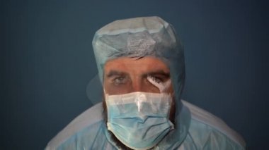 Yorgun sakallı bir doktor sisli bir maske soluyor ve kameraya bakıyor. Yakın plan. Nefes alırken maskeden ter akıyor..