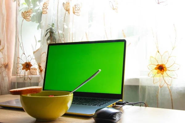 A laptop with a green screen and a plate of food stands on a table in a room near the window — 스톡 사진