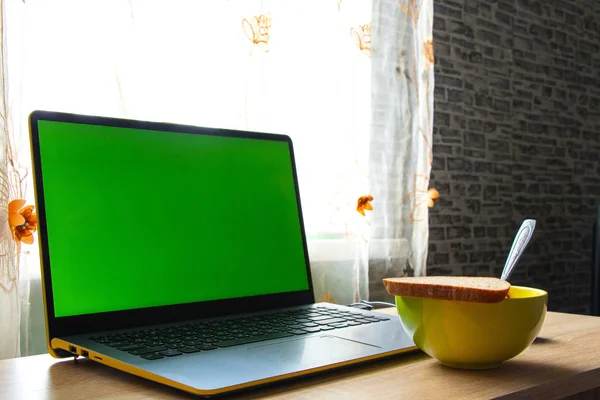 A laptop with a green screen and a plate of food stands on a table in a room near the window — 스톡 사진