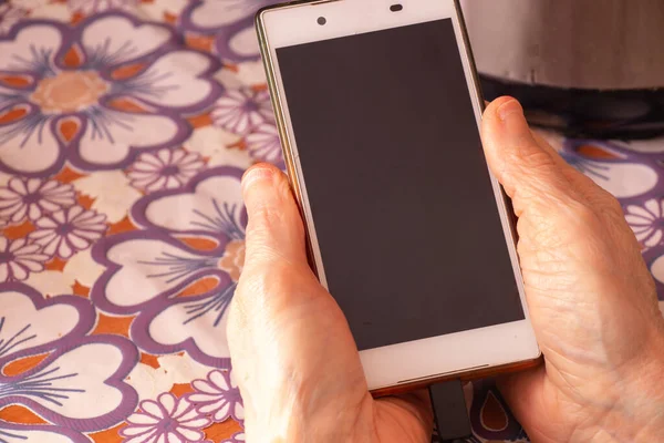 elderly woman holding a phone in her hands while sitting at a table in the kitchen closeup