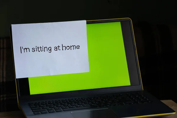 laptop with a green screen on a table in a room at home and a sheet with an inscription in English sitting at home in the period.