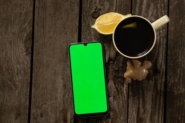 mobile phone with a green screen and a cup of tea with lemon on an wooden table