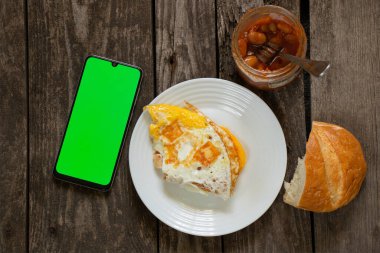 Beyaz bir tabakta kızarmış yumurta, ekmek ve bir kavanoz fasulye ve yeşil ekranlı bir telefon.