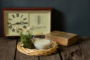 Bir fincan süt ve saat başında papatyalarla dolu bir vazo ve tahta bir masanın üzerinde bir kitap.