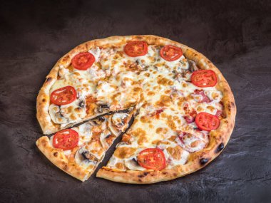 Mantarlı, domatesli ve peynirli pizza koyu bir arka planda yatıyor. Pizzanın kalanının yanında dilimlenmiş bir dilim.