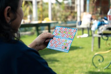 Bir elinde Tambola Housie Loto Bingo bileti olan bir kadın. Bahçede otururken bazı numaralar katlanmış.