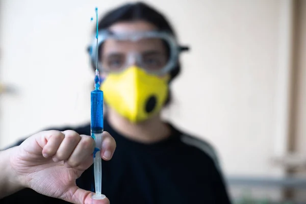 Young indian female healthcare worker doctor nurse holding a syringe with blue vaccine for coronavirus in the current pandemic. The mouth and nose yellow mask and the full cover goggles show the personal protective equipment being used by health prof