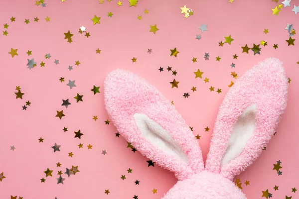 Easter bunny concept. Toy rabbit\'s ears with stars confetti on pastel pink background. Flat lay, top view, copy space