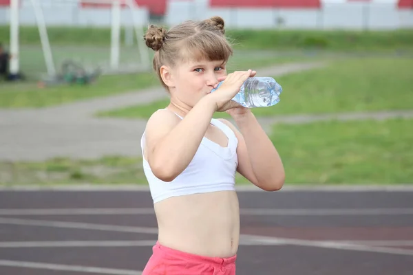 a sporty girl drinks water from a bottle and smiles sweetly at a running stadium