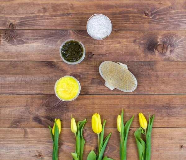 Flatlay view spa cosmetics products scrub and natural brush for face and body on brown wood background with yellow flowers. Top view spa beauty flat lay.