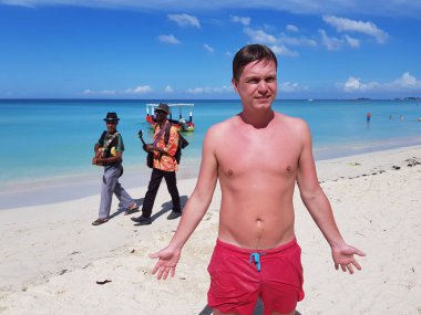 Kumsalda güneşlenirken bir delikanlı, Karayip Denizi, Jamaika, onun arkasında iki müzisyen vardır