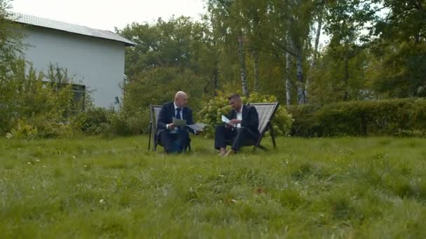 Two men sit in chairs with papers and talk. — Stock Video