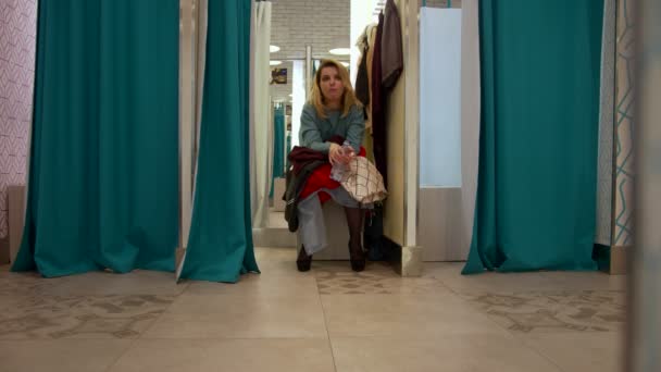 Girl sits in the fitting room with a bunch of clothes — Stock Video