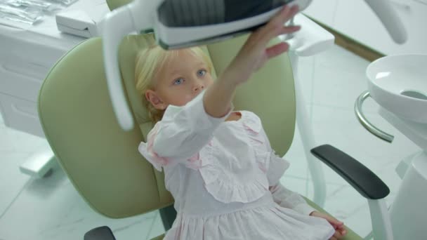 Mała dziewczynka siedzi na fotelu dentystycznym, dotyka lampy — Wideo stockowe