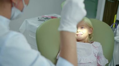Kadın dişçi bir lambayı kızın ağzına doğrultuyor ve dişlerine bakıyor..