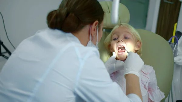 Donna dentista dirige una lampada alla bocca delle ragazze e guarda i suoi denti . — Foto Stock