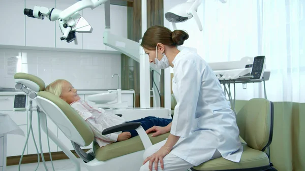 Dentista feminina em máscara fala com uma menina pequena na clínica . — Fotografia de Stock