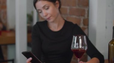 Kadın telefona bakar, birini bekler ve şarap içer.