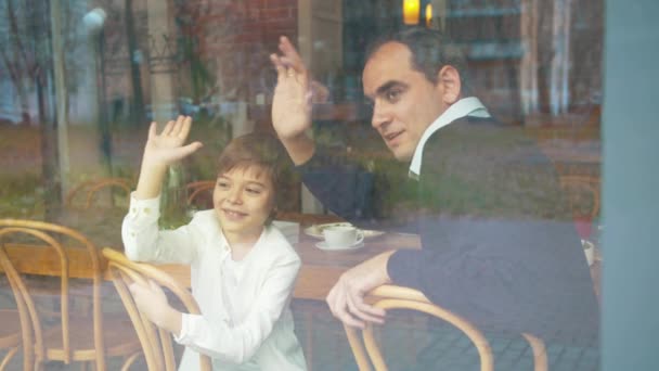 Far och son vinkar till någon genom fönstret i caféet — Stockvideo