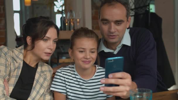 Γονείς και κόρη κοιτάζουν φωτογραφίες στο τηλέφωνο σε ένα καφέ. — Αρχείο Βίντεο