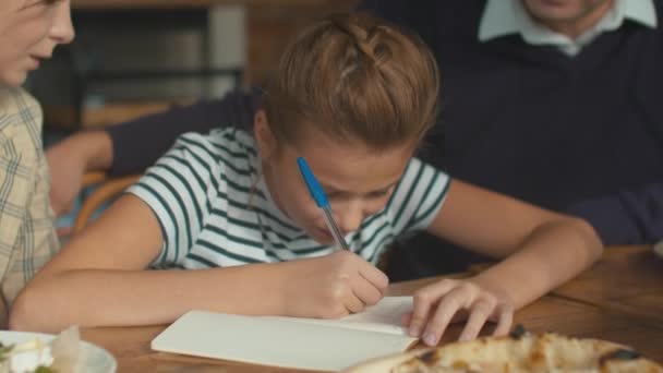 Bella ragazza scrivere qualcosa a pranzo, i genitori si siedono con lei e aiutare — Video Stock