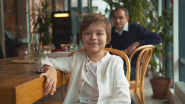 Mały chłopiec siedzi w kawiarni i uśmiecha się — Wideo stockowe