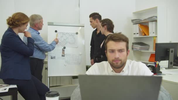 Trött man arbetar på laptop, arbetskamrater dehind honom tittar på system av lägenhet på skrivbordet — Stockvideo