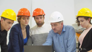 Kask takan iş arkadaşları dizüstü bilgisayarda bir şey gösteren konuşan adamı dinliyorlar. 