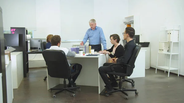 İş arkadaşları masaya oturur, adam gibi durur ve onlara bir şey açıklar.