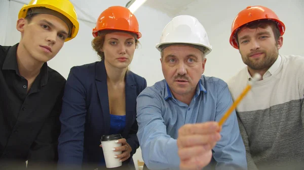 Architect in helmet talks with his workmates and shows a scheme on the screen of laptop