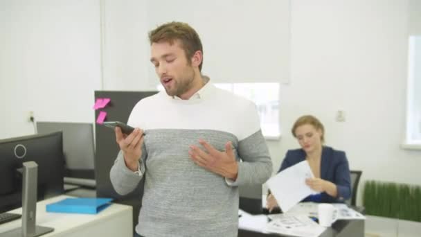 El hombre graba emocionalmente el mensaje de voz en el teléfono, la mujer detrás de él trabaja con dibujos — Vídeo de stock