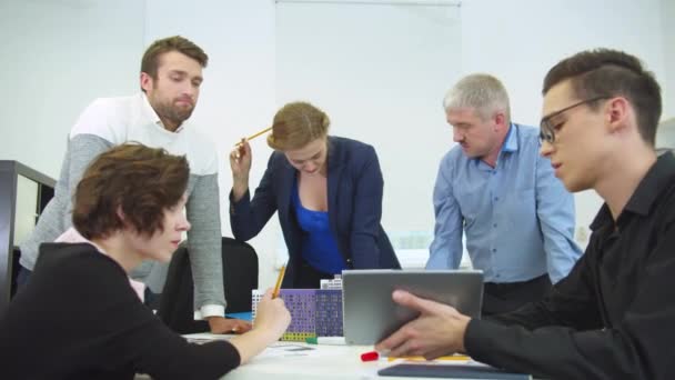 Joven sentado en una mesa con tableta, sus compañeros de trabajo miran y discuten — Vídeo de stock