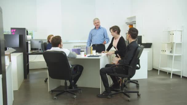 Colegas de trabalho sentam-se em uma mesa, homem levantam-se e parabenizam seu colega — Vídeo de Stock