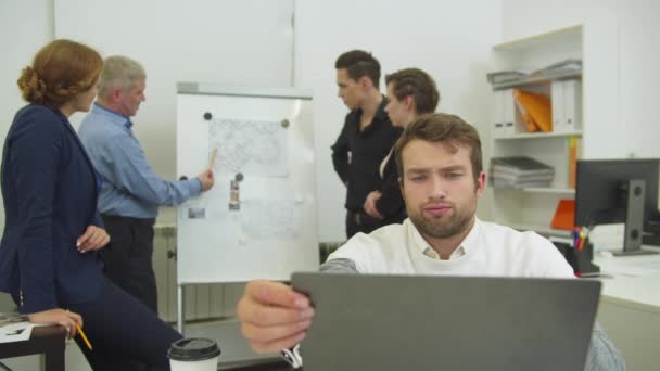 Moe man werkt op de laptop, zijn collega 's bespreken regeling van appartement op bureau — Stockvideo