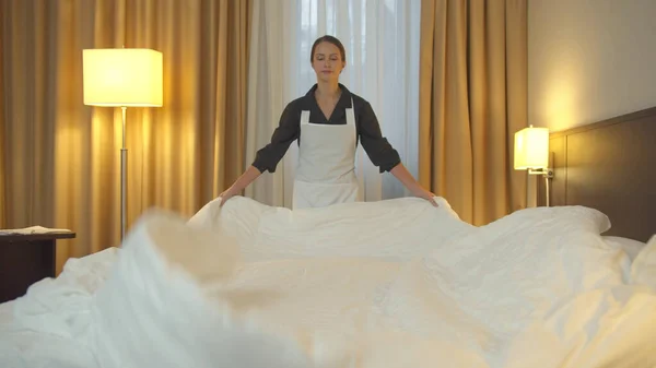 Empregada doméstica em uniforme fazer uma cama e espalhar um cobertor no quarto de hotel — Fotografia de Stock