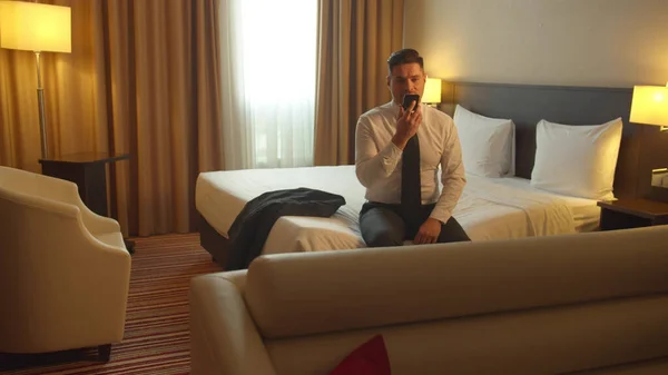 Uomo in camicia e cravatta sedersi sul letto in hotel e registrare il messaggio vocale al telefono — Foto Stock
