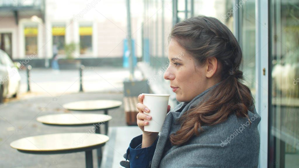 Side view, young woman wraps herself in a grey blanket and drinks coffee